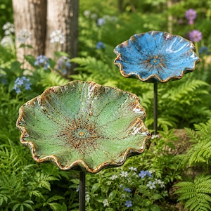 K&ouml;nnte beinhalten: Zwei Gartenstecker aus Keramik mit breiten, gewellten, bl&uuml;tenf&ouml;rmigen Schalen. Eine Schale ist hellgr&uuml;n gesprenkelt mit braunen R&auml;ndern, die andere blau gesprenkelt mit braunen R&auml;ndern. Jede Schale ist auf einem d&uuml;nnen, dunklen Metallstab montiert.