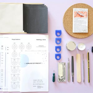 Peut inclure: Un kit de fabrication de bagues DIY avec divers outils et matériaux, dont du papier de verre, des baguiers bleus, un crayon et une brosse à dents. Le kit comprend également un tableau en liège avec un papier "MULTISIZER" et des gabarits.