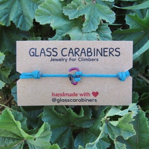 Peut inclure: Un bracelet en cordon bleu turquoise avec un pendentif en forme de mousqueton en verre. Le pendentif est rouge, jaune et bleu. La carte indique "GLASS CARABINERS Bijoux pour grimpeurs fait main avec @glasscarabiners".