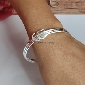May include: A silver bangle bracelet featuring a knot design. The bracelet is composed of several thin, shiny silver bands that encircle the wrist. The text "SilverIndia2022" is visible on the wrist.