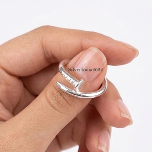 May include: A silver-colored ring in the shape of a nail, with a curved band that wraps around the finger. The nail head features a series of small, rectangular details. The text "SilverIndia2022" is visible on the ring.
