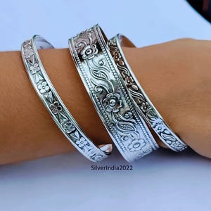 Könnte beinhalten: Vier silberne Armreifen mit floralen und Blattmustern. Die Armreifen werden am Handgelenk getragen und weisen aufwendige Designs auf. Das Metall hat eine helle, reflektierende Oberfläche. Die Armreifen sind eine Art Schmuck.