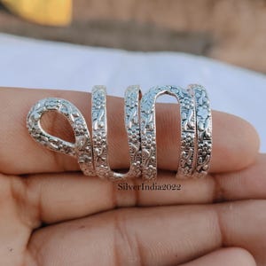 May include: A silver-colored ring with a floral pattern wraps around the finger. The ring has a unique design, with a loop at the top and multiple bands that spiral down the finger. The ring is made of a shiny metal and has a detailed, textured surface.