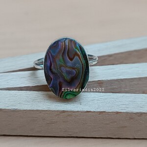 Puede incluir: Un anillo de plata con una incrustación ovalada de concha de abulón. La concha tiene un color iridiscente verde, azul y morado.