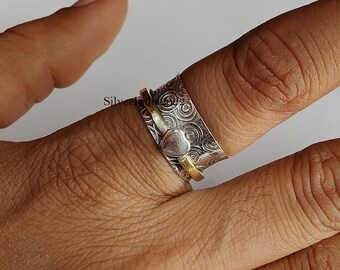 Può includere: Un anello spinner in argento con un accento a forma di cuore. L'anello presenta una fascia strutturata con un motivo a spirale e una fascia color oro. L'accento a forma di cuore è argentato. L'anello è indossato su un dito.
