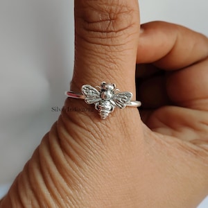 Puede incluir: Anillo plateado con un diseño detallado de abeja. La abeja tiene alas y cuerpo texturizados, con una banda delgada. El anillo se lleva en un dedo, mostrando el diseño de la joya.