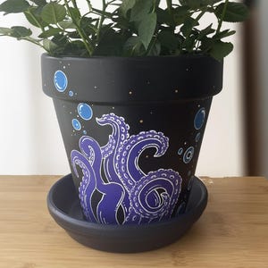 May include: Black ceramic plant pot with a purple octopus design and blue bubble accents. The pot sits on a matching saucer and holds a green plant. The design is hand-painted and adds a unique touch to any home decor.