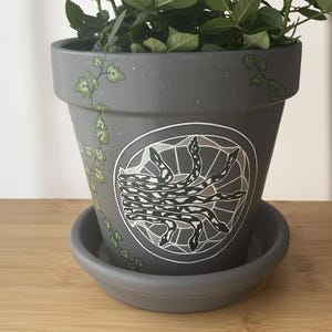 May include: A gray ceramic plant pot with a matching saucer. The pot is decorated with a white and black geometric design featuring snakes. Green vines and leaves are painted around the rim. The pot is filled with green plants.