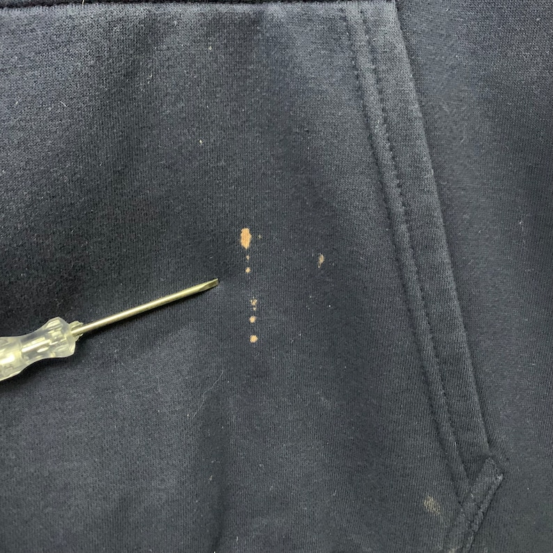 May include: Close-up of a navy blue hoodie pocket with brown stains. A silver screwdriver is pointing at the stains. The hoodie's fabric is slightly textured.