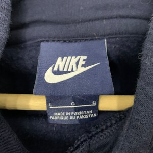 May include: A close-up of a navy blue Nike hoodie label. The label displays the Nike logo in white and the text "MADE IN PAKISTAN" and "FABRIQUE AU PAKISTAN". Size indicators are also visible.