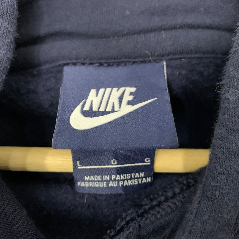 May include: A close-up of a navy blue Nike hoodie label. The label displays the Nike logo in white and the text "MADE IN PAKISTAN" and "FABRIQUE AU PAKISTAN". Size indicators are also visible.