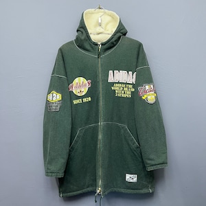 Może przedstawiać: Zielona kurtka z kapturem Adidas z kremową podszewką z polaru. Kurtka posiada wiele logo i naszywek z tekstem Adidas, w tym "Adidas Since 1920" i "Adidas The World Brand With The 3 Stripes".