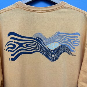 May include: Mustard-colored t-shirt with a wavy graphic design in shades of blue. The graphic includes the text "JUST JUMP" in a light blue color. The shirt has a crew neck and short sleeves.