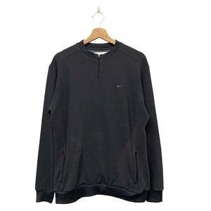 May include: Black Nike sweatshirt with a quarter-zip neckline and a small Nike logo on the chest. The long-sleeved sweatshirt has two side pockets and a ribbed hem and cuffs. The garment is hanging on a wooden hanger.