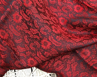 Könnte beinhalten: Nahaufnahme eines tiefroten Stoffes mit floralem und Paisley-Muster. Das Design zeigt detaillierte rote Blumen und Blätter vor einem dunklen Hintergrund. Der Stoff ist drapiert und zeigt seine Textur und Falten. Eine weiß-schwarz gemusterte Oberfläche ist unten sichtbar.