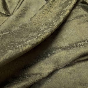 Op de afbeelding: Close-up van een textuurrijke, groene stof met een subtiel bloemenpatroon. De stof is zacht en drapeert mooi.