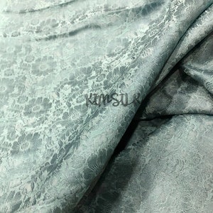 Könnte beinhalten: Nahaufnahme eines hellblauen Stoffes mit einem dezenten Blumenmuster. Der Stoff ist in der Mitte gefaltet und zeigt die Textur und den Glanz des Materials.