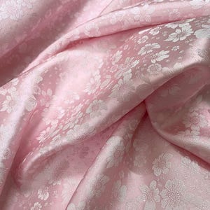 Könnte beinhalten: Rosa Satin-Stoff mit einem weißen Blumenmuster. Der Stoff ist weich und luxuriös, und das Blumenmuster ist zart und feminin.