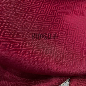 May include: Close-up of a deep red fabric with a repeating geometric pattern of squares and lines. The fabric has a subtle sheen and a textured surface.