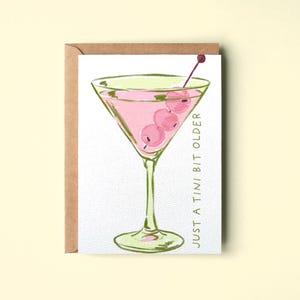 Op de afbeelding: Een wenskaart met een aquarelillustratie van een martiniglas gevuld met een roze cocktail en drie olijven aan een prikker. De tekst luidt JUST A TINI BIT OLDER in groene hoofdletters. De kaart zit in een bruine kraftpapieren envelop.