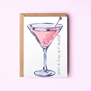 Op de afbeelding: Een wenskaart met een aquarelillustratie van een martiniglas gevuld met een roze cocktail en drie olijven op een prikker. De tekst "JUST A TINI BIT OLDER" is verticaal in zwarte inkt gedrukt aan de rechterkant van de witte kaart.