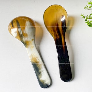 May include: Two handcrafted horn spoons. One spoon features a light brown and white marbled pattern, while the other has a dark brown and amber color gradient. The spoons are displayed on a white surface.