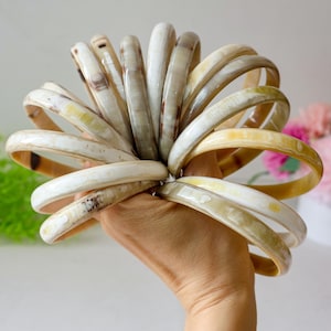 May include: A collection of cream-colored bangles with brown and yellow streaks. The bracelets are held in a hand, showcasing their smooth, polished surfaces. The bangles are made of a natural material, and the color variations add to their unique appeal.
