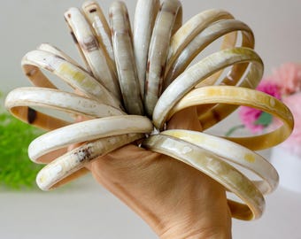 Puede incluir: Una colección de brazaletes de color crema con vetas marrones y amarillas. Los brazaletes se sostienen en una mano, mostrando sus superficies lisas y pulidas. Los brazaletes están hechos de un material natural, y las variaciones de color se suman a su atractivo único.