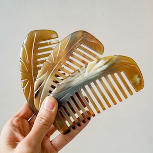 Peut inclure: Trois peignes artisanaux en matériau brun clair, peut-être en corne, sont présentés. Chaque peigne présente un motif de feuille sculptée sur le manche et des dents espacées. Les peignes sont tenus dans une main sur un fond neutre, mettant en valeur leurs variations de couleur naturelles.