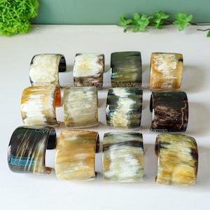 Puede incluir: Una colección de 12 pulseras de cuerno en varios tonos de marrón, beige y negro. Las pulseras están apiladas en tres filas de cuatro, con cada pulsera teniendo un patrón único.