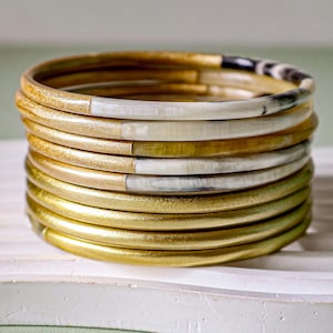 May include: A collection of gold and ivory bangles. The bangles are smooth and rounded, with a metallic gold finish. Some bangles feature ivory or patterned sections. The stack is presented on a white surface.