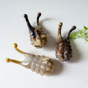 May include: Three carved snail figurines with textured bodies and curved antennae. The snails are in shades of brown and tan, with a glossy finish. The figurines are displayed on a white surface.