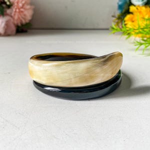 May include: Two stacked bangles. The top bangle is a light tan color with dark brown accents. The bottom bangle is black. The bangles are smooth and curved, with a glossy finish. The background is blurred, with hints of flowers.