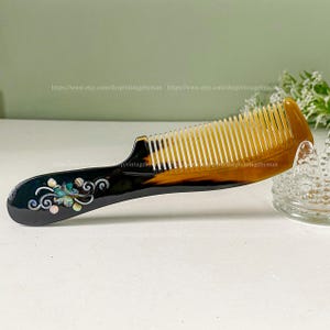 May include: A brown and black tortoiseshell comb with a decorative handle featuring a floral design with iridescent accents. The comb has fine teeth and is made of natural materials.