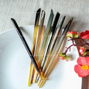 Può includere: Una collezione di forcine per capelli fatte a mano in varie tonalità di marrone, nero e crema. Gli accessori per capelli sono disposti su un piatto bianco, mostrando la loro finitura lucida e le variazioni naturali. Le punte sono appuntite e i bastoncini hanno un design liscio ed elegante.
