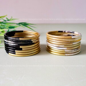 Può includere: Una pila di 10 braccialetti dorati con accenti neri e bianchi. I braccialetti sono realizzati in un materiale liscio e lucido e hanno un design semplice ed elegante.