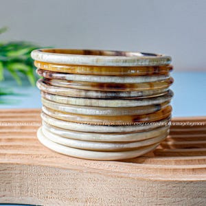 Peut inclure: Une pile de 12 bracelets en corne d'ivoire et brune. Les bracelets sont empilés sur une surface en bois.