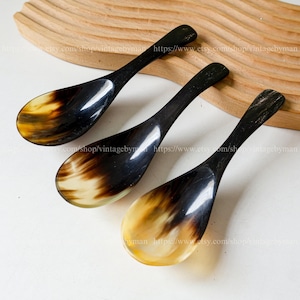 May include: Three small spoons made of dark brown and light yellow horn. The spoons are arranged in a row on a wooden surface.