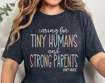 May include: Dark gray t-shirt with the text "caring for TINY HUMANS and STRONG PARENTS" in pastel colors. The shirt also includes the hashtag "#NFP NURSE". The shirt is a crew neck style.