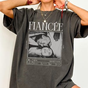 May include: Dark gray oversized t-shirt with the word "FIANCÉE" in large white letters. Below the word is a black and white photo of a couple. The shirt also has the words "ENGAGED TO DAVID".