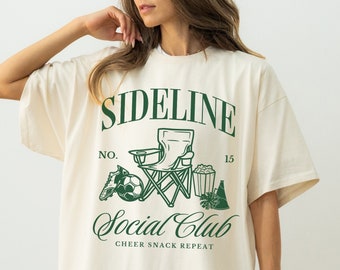 May include: An oversized white t-shirt with a green graphic print that says "Sideline Social Club" with a chair, football, popcorn, and a party hat. The text below the graphic says "Cheer Snack Repeat". The shirt is labelled "Ivory".