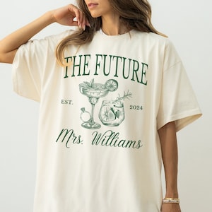 May include: Ivory t-shirt with green text that reads "The Future Est. 2024 Mrs. Williams" with a drawing of two cocktails.
