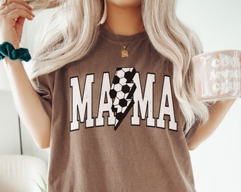 May include: A brown t-shirt with the word "MAMA" in white letters with a lightning bolt design featuring a football inside. The shirt is being worn by a person.