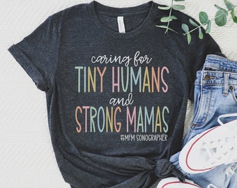 May include: Dark gray t-shirt with the text "caring for tiny humans and strong mamas" in pastel colors. The shirt is styled with blue jeans and white sneakers. A sprig of greenery is placed on the top right.