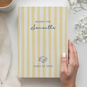May include: A hardcover book with a yellow and white vertical striped cover. The text reads WISHES FOR Samantha and CLASS OF 2026, accompanied by a blue line-art icon of a graduation cap and diploma centered near the bottom of the front cover.