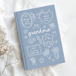 May include: A light blue hardcover book featuring white line art illustrations of flowers, a teacup, and frames. The text reads blessed and loved, olivia, jones, max, grandma, mom EST. 1995, grandma EST. 2025, and the best chapter yet.