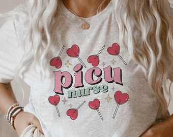 Puede incluir: Una camiseta gris con el texto "picu nurse" en letras rosas y verdes. El texto está rodeado de corazones rosas en palos de piruleta.