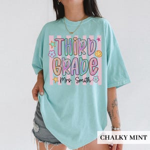 May include: A chalky mint green t-shirt featuring a graphic with vertical pink stripes and the text "THIRD GRADE" in colorful, bubble-style lettering. Below the text, "Mrs. Smith" is written in black script, surrounded by small star and flower icons.
