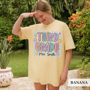 May include: A pale yellow short-sleeve t-shirt featuring a graphic print with light blue vertical stripes. The text reads "THIRD GRADE" in colorful bubble letters, followed by "Mrs. Smith" in black script, surrounded by stars and flowers.