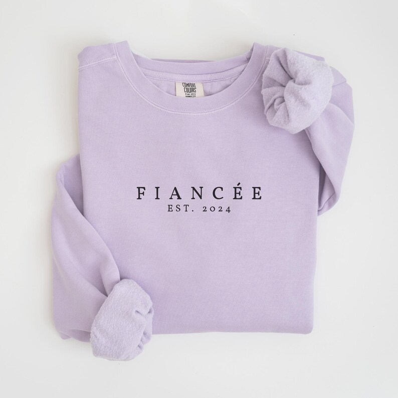 May include: A lavender sweatshirt with the text "FIANC&Eacute;&Eacute; EST. 2024" printed on the front.